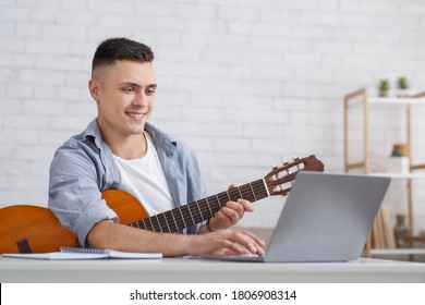 Online Music Lesson During Covid-19 Pandemic. Smiling Young Man With Guitar Looking At Laptop In Living Room Interior, Free Space