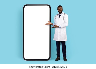 Online Offer. Black Smiling Doctor In White Robe Pointing At Big Blank Smartphone With Empty Screen For Mockup, African American Therapist Showing Copy Space For Mobile App Design Or Advertisement