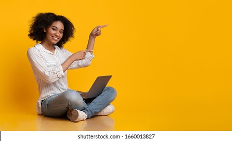 Online Offer. Cheerful Afro Girl Sitting On Floor With Laptop And Pointing Away With Fingers, Indicating Copy Space On Yellow Background, Panorama