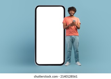 Online Offer Concept. Happy Curly Young Indian Man Using Cell Phone On Blue Studio Background, Standing By Big Mobile Phone With White Empty Screen, Full Length Shot, Panorama With Copy Space, Mockup