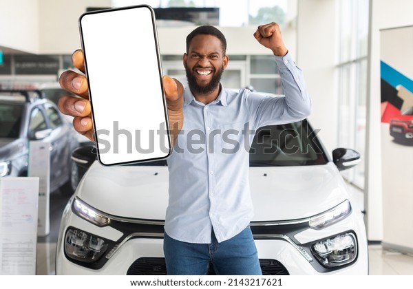 Online Offer. Excited Black Man With Blank\
Smartphone Standing In Car Showroom, Happy Emotional African\
American Male Demonstrating Cellphone With White Screen At Camera\
And Raising Fist, Mockup