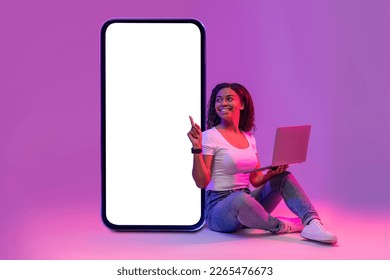 Online Offer. Happy black woman with laptop pointing at big blank smartphone with white screen while sitting in neon light over purple studio background, smiling lady recommending new app, mockup