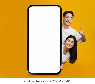 Online Offer. Smiling Asian Couple Peeking Out Behind Big Blank Smartphone And Showing Thumb Up Over Yellow Background, Happy Young Man And Woman Recommending New Mobile App Or Website, Mockup