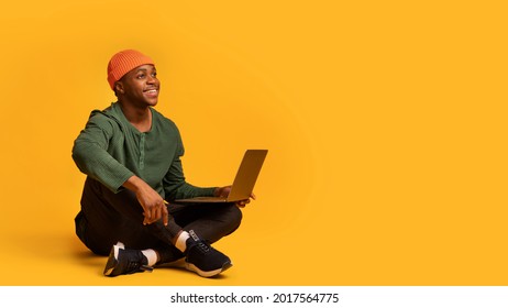 Online Offer. Smiling Young Black Guy Sitting With Laptop On Floor And Looking Away At Copy Space On Yellow Background, Portrait Of Dreamy Millennial African American Man With Computer, Panorama