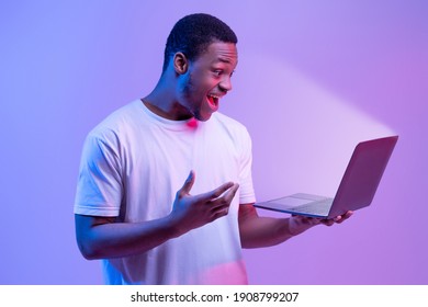 Online Offer. Surprised African American Man Holding Laptop Looking At Glowing Screen, Excited Black Guy Playing Games Or Browsing Internet On Computer, Stranding In Neon Light Over Purple Background