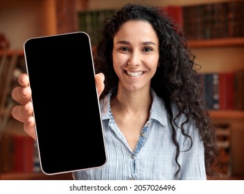 Online Offer. Young Smiling Lady Showing Blank Black Smartphone Screen At Camera, Millennial Female Recommending New Edcuational Website Or Mobile Application While Sitting In Library, Mockup