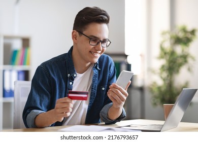 Online Order. Smiling Young Male Entrepreneur Using Smartphone And Credit Card While Sitting At Desk In Office, Happy Millennial Man Shopping In Internet, Enjoying E-Commerce, Copy Space