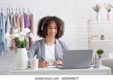 Online Orders For Small Businesses During Covid-19 Quarantine. Smiling Business Woman In Headphones Working With Clients Or Watching Webinar In Laptop In Living Room Interior With Rack With Clothes