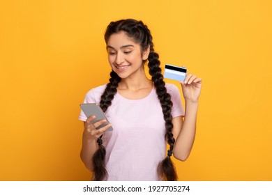 Online Payment. Frontal Portrait Of Smiling Indian Woman Holding And Showing Debit Credit Card, Using Her Smartphone For Shopping Posing Isolated Over Yellow Studio Background. Money Transfer