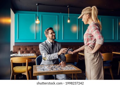 Online Payment Of Restaurant Bills. Man In A Business Suit With A Tie Sits At A Table In A Restaurant And Leans His Phone On The Terminal Held By A Female Waitress. Paying For A Meal During Lunch Time