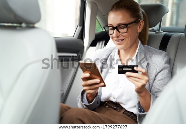 Online payment. Smiling business woman\
wearing eyeglasses using her smartphone and credit card to buy\
something while sitting on back seat in the\
car