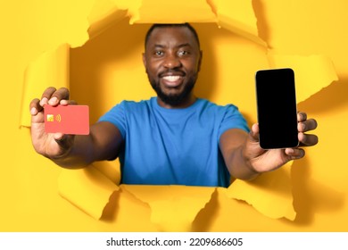 Online Payments. Happy African American Man Holding Blank Smartphone And Credit Card, Standing Breaking Through Yellow Paper Background, Recommending Mobile App Or Website, Mockup