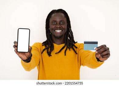 Online Payments. Smiling Black Guy Holding Blank Smartphone And Credit Card While Standing On White Studio Background, Cheerful African American Man Recommending Mobile App Or Website, Mockup