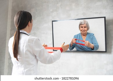 Online physiotherapy for the elderly. Doctor showing an elderly woman exercises with a fitness rubber band. Remote medicine during quarantine. Female doctor consults on video conference. - Shutterstock ID 1822781096