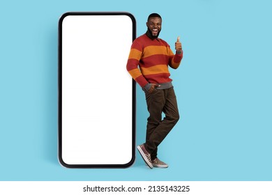 Online Promo. Joyful Black Man Standing Near Big Blank Smartphone And Showing Thumb Up, Cheerful African American Male Leaning At Mobile Phone With White Screen For Mockup, Full Length Shot - Shutterstock ID 2135143225