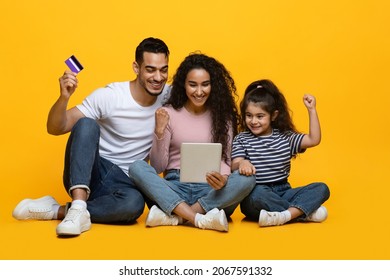 Online Sales. Cheerful Middle Eastern Family Of Three Using Digital Tablet And Credit Card, Celebrating Success While Making Internet Shopping, Enjoying Big Discounts, Sitting Over Yellow Background