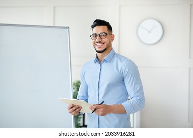 Online Schooling. Positive Arab Male Teacher Standing Near Empty Blackboard With Clipboard, Giving Remote Lecture On Web, Speaking To Students, Mockup. Happy Young Tutor Conducting Lesson On Internet