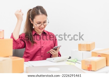 Online selling concept, happy and smiling Asian woman, reading message on smartphone screen, desk with laptop Cardboard boxes and calculators.