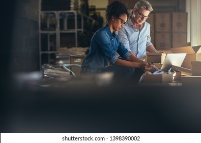 Online Shop Business Partners Working In Small Office Together. Woman Packing The Purchased Product In The Box With Man Updating The Shipment Status On Laptop.