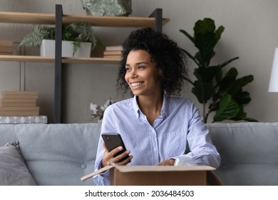 Online shopper. Smiling afro american woman web shop client sit on sofa open parcel with consumer goods make easy payment for delivery by phone. Young black lady hold cell shoot mail package unboxing