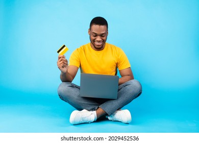 Online Shopping. African American Man Using Laptop Holding Credit Card Purchasing Things In Internet Shop Sitting Over Blue Studio Background. E-Commerce Technologies Concept