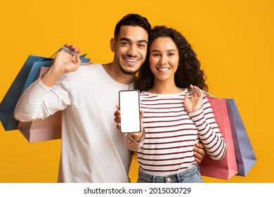 Online Shopping App. Cheerful Young Arab Couple Showing Smartphone With Blank White Screen At Camera And Holding Bright Shopper Bags While Standing Isolated Over Yellow Studio Background, Mockup