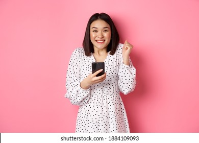 Online Shopping And Beauty Concept. Excited Asian Woman Winning In Internet, Holding Smartphone And Rejoicing, Smiling Happy And Celebrating, Standing Over Pink Background