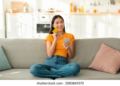 Online Shopping Concept. Dreamy Asian Woman With Smartphone And Credit Card Sitting On Couch, Thinking About Internet Purchases Or Food Delivery, Free Space