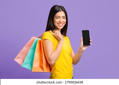 Online Shopping Concept. Happy Asian Girl With Shopper Bags Demonstrating Black Smartphone Screen, Mockup Image With Copy Space