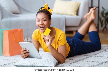 Online Shopping Concept. Happy Black Woman Using Digital Tablet, Holding And Showing Credit Card, Buying And Ordering Things In Internet Shop Lying On The Floor Carpet, Looking At Camera