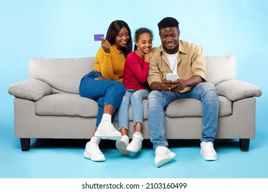 Online Shopping Concept. Happy Young Black Parents With Daughter Using Cell Phone And Debit Credit Card Buying Goods In Internet Web Store, Three People Sitting On Couch Isolated On Blue Studio Wall