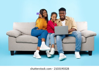 Online Shopping Concept. Happy Young Black Parents With Daughter Using Laptop And Debit Credit Card For Buying Goods In Internet Web Store, Three People Sitting On Couch Isolated On Blue Studio Wall