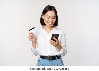 Online Shopping Concept. Korean Woman Holding Credit Card And Looking At Smartphone App, Buying, Order Delivery In Mobile Phone Application, Standing Over White Background