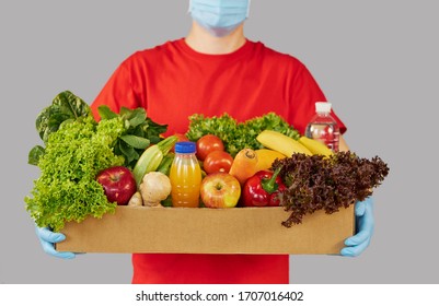 Online Shopping Concept. Male Courier In Red Uniform, Protective Mask And Gloves With A Grocery Box With Fresh Fruits And Vegetables. Home Delivery Food During Quarantine Coronavirus 