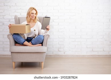 online shopping concept - portrait of young beautiful woman sitting in armchair with tablet pc and cardboard box - copy space over white brick wall background