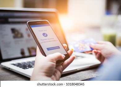 Online shopping concept, young woman hands holding mobile phone showing payment success information on screen with credit card and laptop computer on table while relax at home