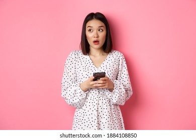 Online Shopping. Confused Asian Girl Holding Smartphone, Looking Clueless At Upper Left Corner, Standing Over Pink Background