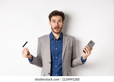 Online Shopping. Confused Man In Suit Spread Hands Sideways, Holding Plastic Credit Card With Mobile Phone And Shrugging Cueless, Standing On White Background