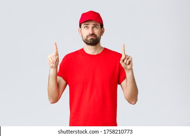 Online Shopping, Delivery During Quarantine And Takeaway Concept. Displeased, Reluctant Young Courier, Employee In Red Uniform Cap And T-shirt, Smirk Uncertain, Reading Bad News Upwards, Point Up