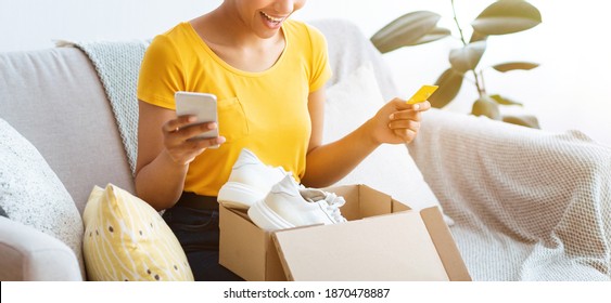 Online Shopping And Delivery Service Concept. Unrecognizable black woman holding credit card and cellphone, received package, unpacking cardboard box with white sneakers, banner, panorama