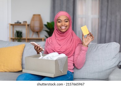 Online shopping and delivery service concept. Excited muslim lady holding credit card, cellphone and received package, unpacking cardboard box, sitting on sofa and smiling at camera
