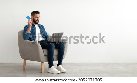 Online shopping, e-commerce, remote banking. Young Arab man sitting in armchair with laptop, using credit card to purchase goods on web, buying products in internet store, copy space. Banner design