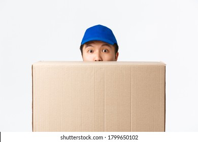 Online shopping, fast shipping concept. Surprised cute asian male courier, delivery man in blue cap, peeking behind large box with client order, bring package to customer doorstep, white background