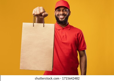 Online Shopping And Grocery Delivery Service. African Courier Giving Shopper Bag With Food Smiling To Camera On Yellow Studio Background.