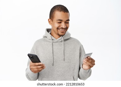 Online Shopping. Handsome Smiling Guy Enter Credit Card Info On Mobile Phone, Paying With Smartphone App, Order In Internet, Standing Over White Background