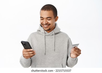 Online Shopping. Handsome Smiling Guy Enter Credit Card Info On Mobile Phone, Paying With Smartphone App, Order In Internet, Standing Over White Background