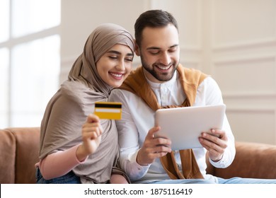 Online Shopping. Happy Muslim Couple Using Digital Tablet And Credit Card Buying And Purchasing Things Sitting On Sofa At Home. Joyful Customers, Modern Shopping And Sales Concept