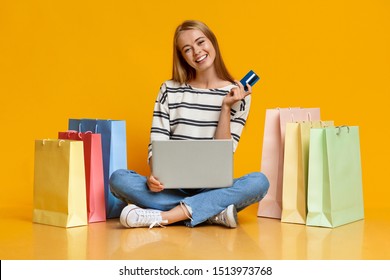 Online Shopping. Joyful Girl Sitting With Laptop And Credit Card Surrounded By Bright Paper Bags Over Orange Studio Background