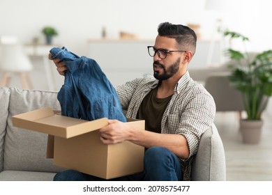 Online Shopping Mistake, Terrible Purchase, Wrong Delivery Concept. Unhappy Young Arab Guy Opening Carton Package, Receiving Clothes, Dissatisfied With Delivered Item At Home