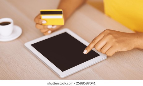 Online Shopping And Payment Concept. Unrecognizable black woman holding credit card and using digital tablet with empty screen for mockup, lady sitting at desk, blurred background, selective focus